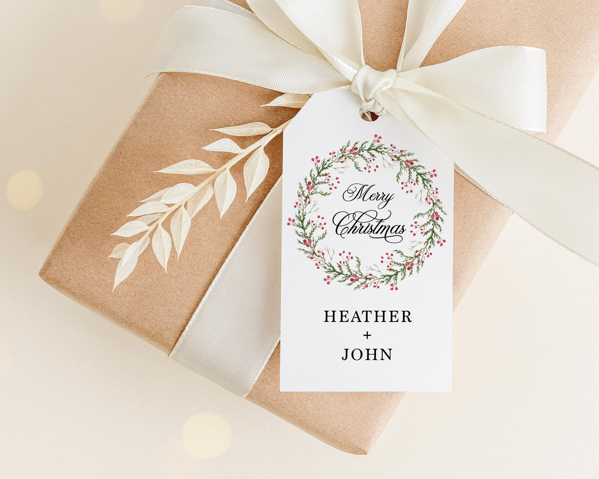 A brown gift box with a white ribbon and a white tag attached to it, featuring a wreath design and the text "Merry Christmas" and "Heather + John".
