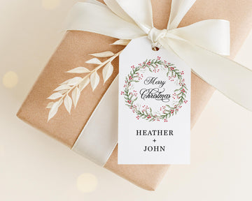 A brown gift box with a white ribbon and a white tag attached to it, featuring a wreath design and the text "Merry Christmas" and "Heather + John".