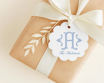 A brown gift box with a white ribbon and a white tag featuring a monogrammed logo and text.