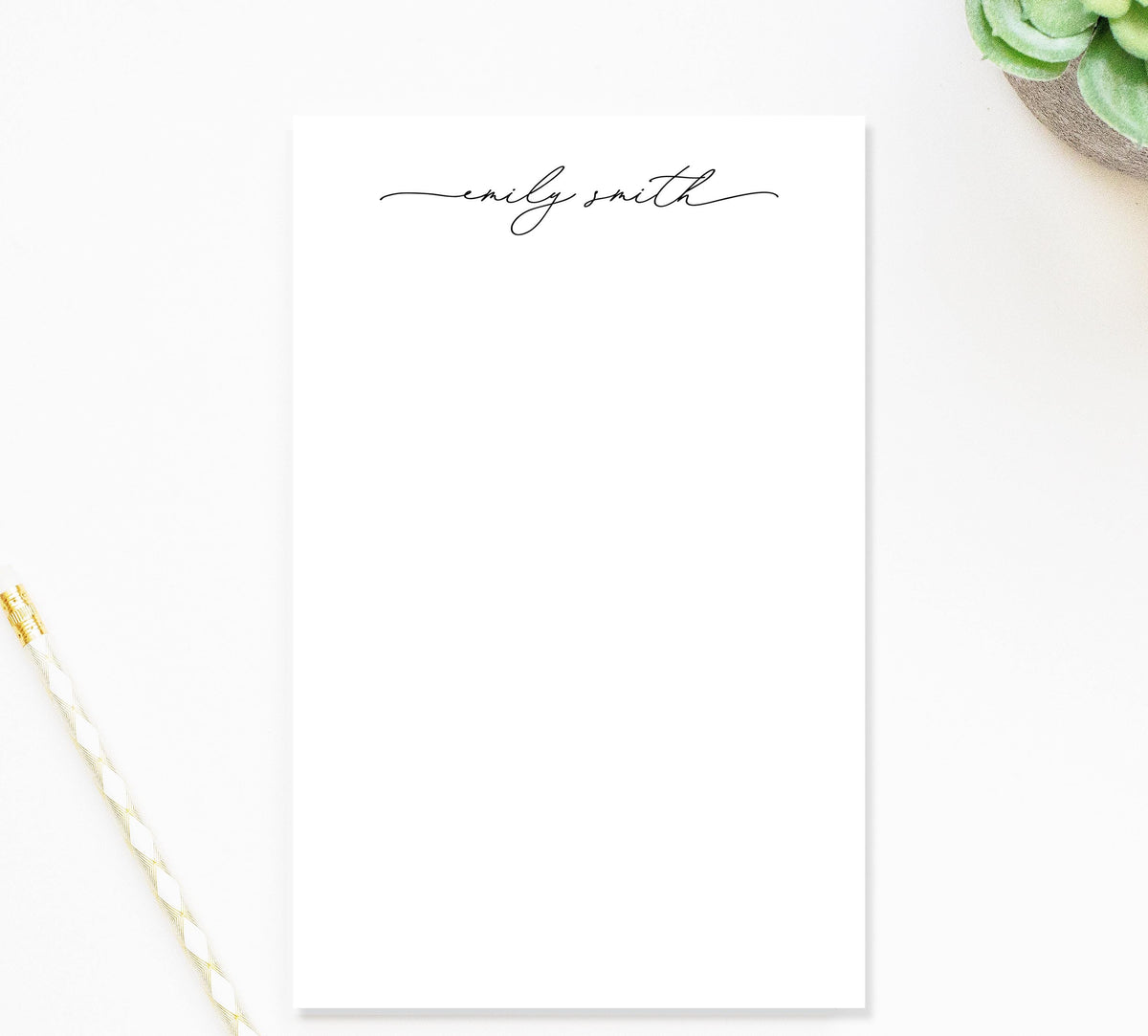a blank white notepad with the name 'Emily Smith' written on it in a cursive font. The notepad is placed on a white surface, and there is a pen and a plant nearby.