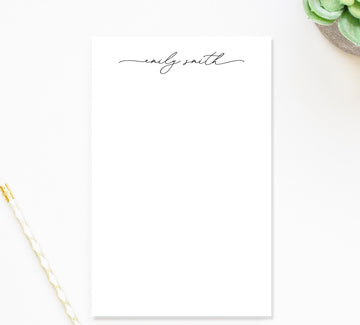 a blank white notepad with the name 'Emily Smith' written on it in a cursive font. The notepad is placed on a white surface, and there is a pen and a plant nearby.