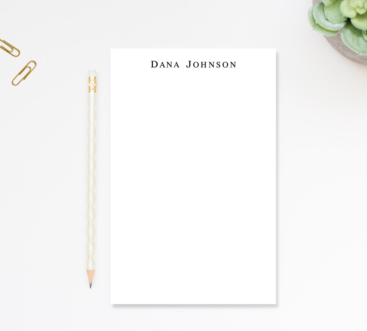 a blank white notepad with the name 'Dana Johnson' written on it, accompanied by a pencil and a small plant.