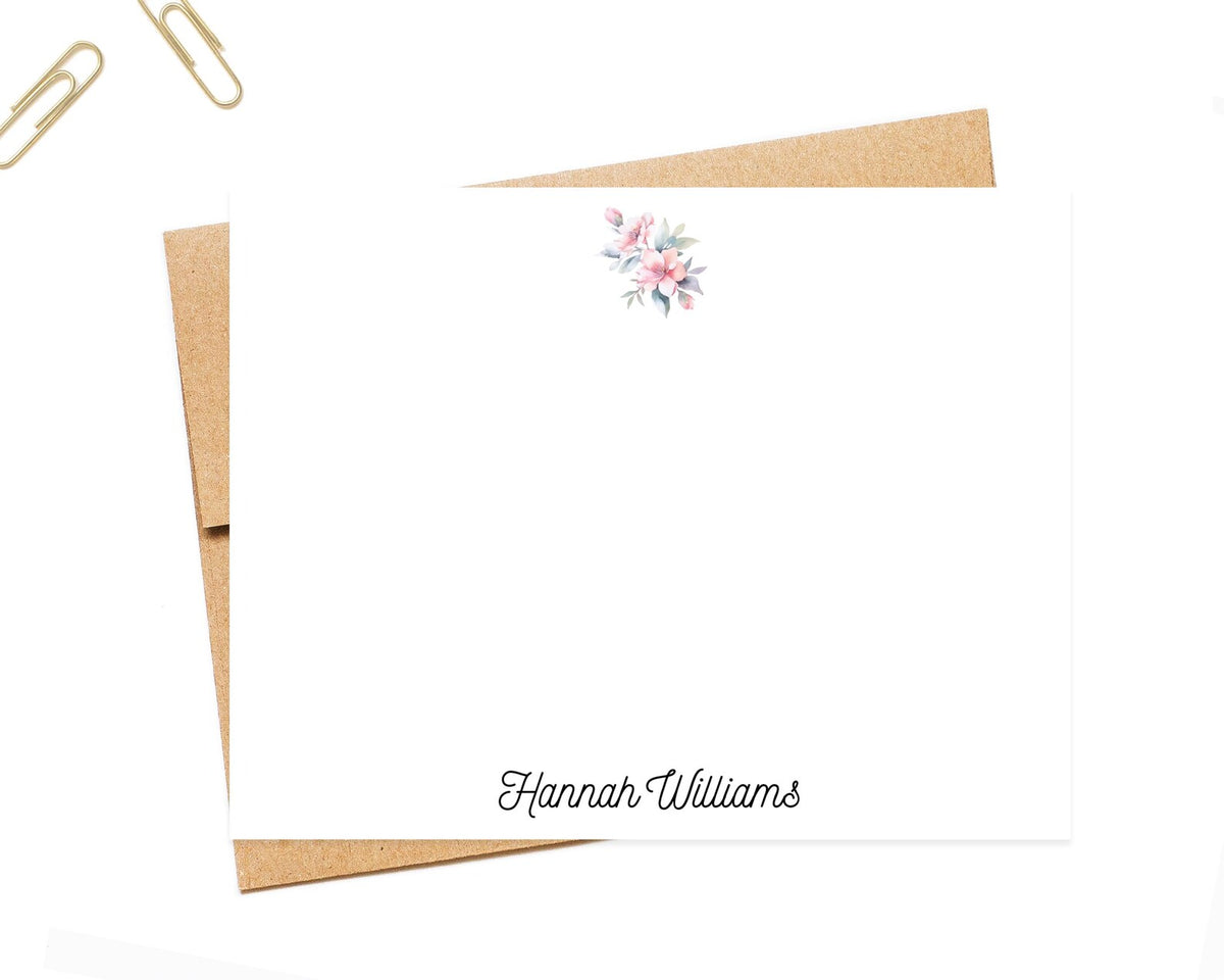 a blank card with a floral design and a name printed on it, along with a brown envelope and a pair of gold paper clips.