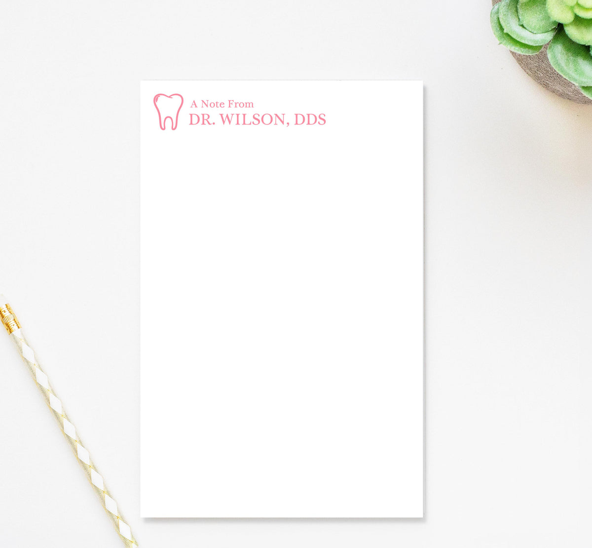 A blank white notepad with a tooth logo and text on it, placed on a white surface next to a pen and a small potted plant.