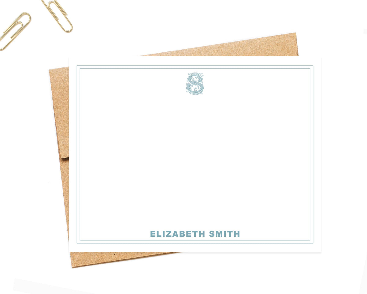 a blank personalized note card with the name 'ELIZABETH SMITH' printed on it, along with a decorative monogrammed 'S' above the card. The card is placed on a white surface, with a pair of gold-colored paper clips visible in the top left corner.