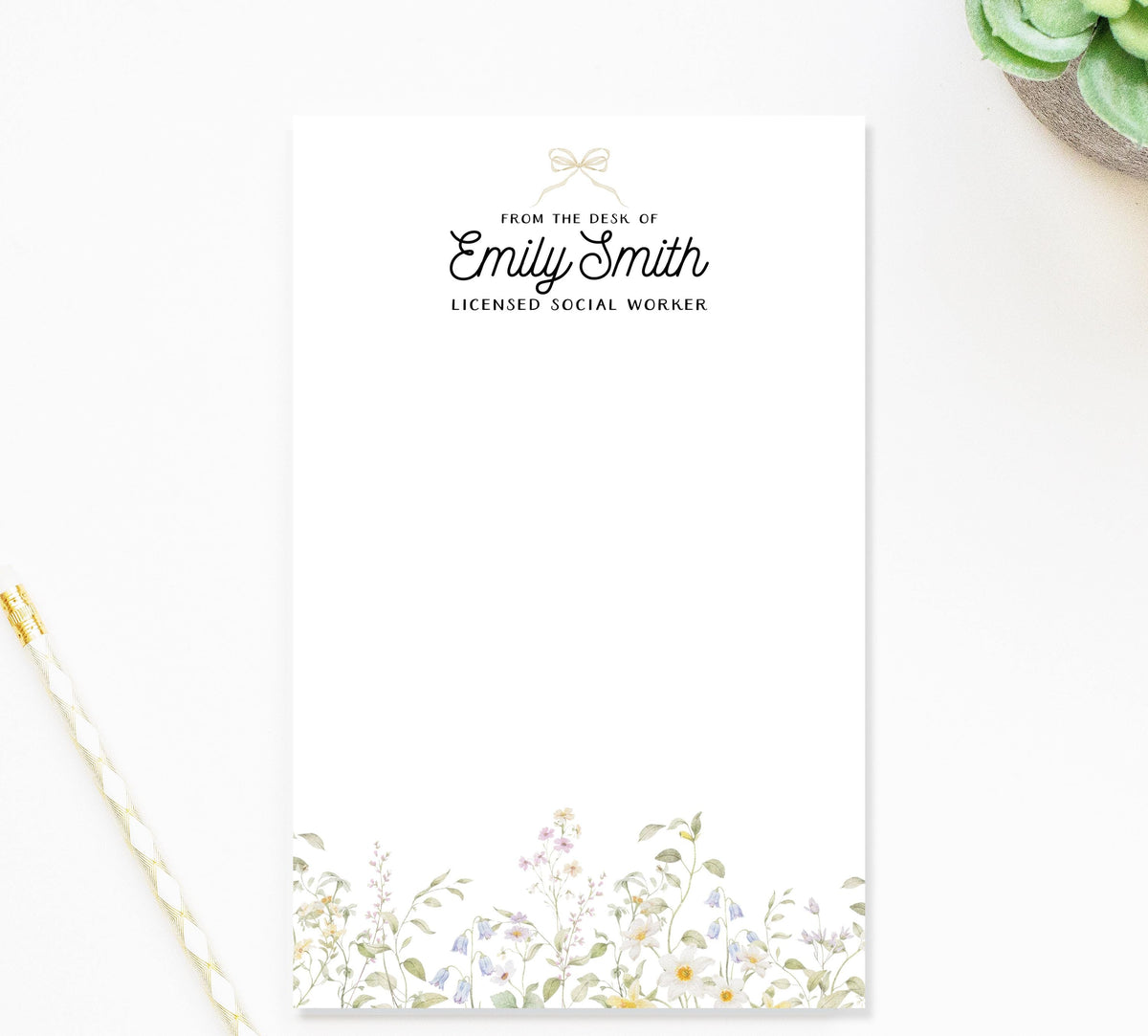 a blank white note card with a floral border, placed on a white surface next to a pen and a small green plant.