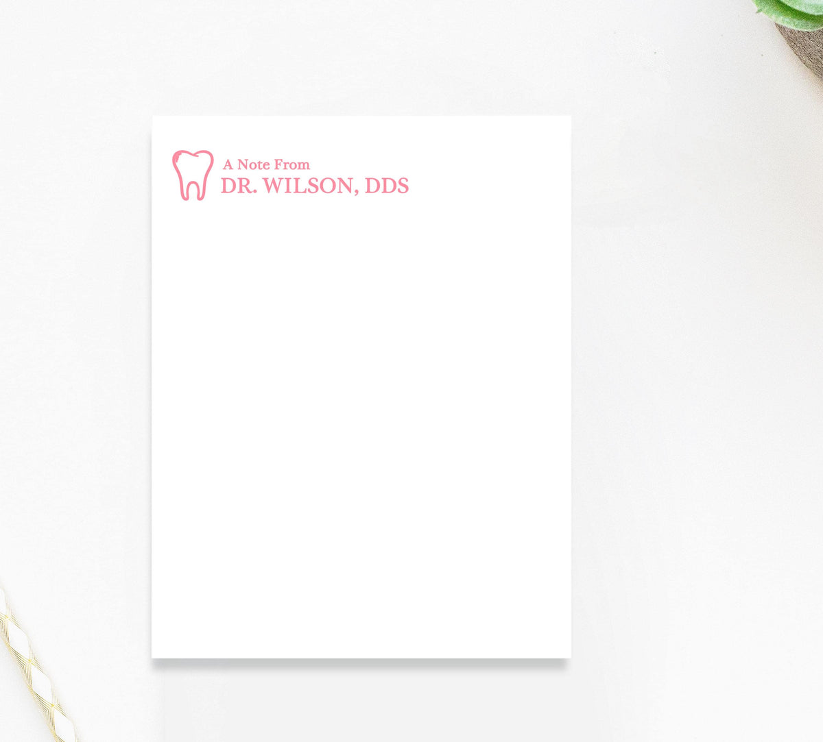 A blank white notepad with a pink logo and text on it, placed on a white surface next to a plant.