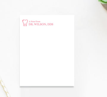 A blank white notepad with a pink logo and text on it, placed on a white surface next to a plant.